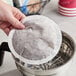 A hand holding a round white Maxwell House coffee filter over a metal coffee maker.