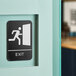 A Lavex black and white plastic exit sign with Braille on a wall above a door with a person running out.