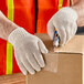 A person wearing Lavex natural cotton and polyester work gloves cuts a box with a knife.