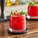 A glass of red strawberry drink with ice, basil, and strawberries.