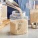 A person pouring white grains into a clear square PET jar.