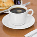 A Libbey white porcelain tea saucer with a cup of coffee on a table with a croissant and jam.