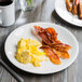A Libbey Royal Rideau white coupe plate with scrambled eggs and bacon on a table.