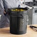 A woman in gloves holding a BWAY black round pail with metal handle full of food.