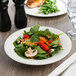 A Libbey Royal Rideau white porcelain bowl filled with salad, spinach, and mushrooms on a table with a fork on a napkin.