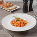 A white Libbey porcelain bowl filled with pasta topped with a sprig of rosemary on a table with a fork and a glass of wine.