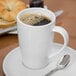 A white porcelain mug filled with coffee, placed on a white saucer with a spoon.