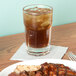 A Libbey Gibraltar highball glass of brown liquid with ice on a table with food.