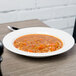 A Libbey Royal Rideau white porcelain deep rimmed soup bowl on a table with a bowl of soup and a spoon.