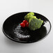 A black Elite Global Solutions round plate with broccoli and tomatoes on it.