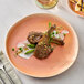 A round primrose peach coupe china plate featuring two lamb chops with garnishes, displayed on a marble surface.