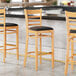 Three Lancaster Table & Seating natural finish wood bar stools with black vinyl cushions next to a counter.