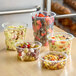 A set of clear round recycled PET plastic deli containers filled with various salads and fruits.