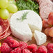 A piece of Cypress Grove PsycheDillic goat cheese on a wooden surface with a slice cut out, grapes, and other fruit.