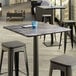 A Lancaster Table & Seating square table top with a smooth finish on a table with stools and a blue glass on it.