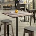 A Lancaster Table & Seating square table top with a textured wooden finish on a table with stools and a glass on it.