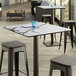 A Lancaster Table & Seating Excalibur square table top with a smooth finish on a table with stools and a glass on it.