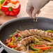 A hand wearing a glove sprinkles ground fennel seed over sliced sausage and bell peppers in a frying pan.