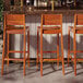 Three Lancaster Table & Seating Virelli Tigerwood slat back barstools sitting in front of a bar.
