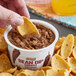 A person holding a chip and dipping it into a yellow container of Furmano's Chipotle Refried Bean Dip.