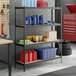 A black metal shelving unit with four particle board shelves, holding various storage containers and supplies in a garage setting.