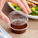 A hand holding a Choice clear plastic souffle container with brown liquid and a lid.