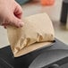 A hand pulling a OneUp by Choice Kraft Premium 2-Ply interfolded napkin from a dispenser on a counter.