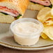 A plastic container of dip next to a sandwich on a plate.