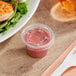 A plastic container of food with a clear plastic lid next to a plate of food with a white spoon and a cup of pink sauce.