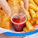 A hand holding a Choice clear plastic souffle cup with red sauce on a plate of chicken nuggets.