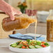 A hand pouring salad into a clear glass juice bottle.