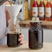 A person pouring dark liquid into a clear glass bottle with a white cap.