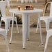 A Lancaster Table & Seating white metal outdoor table on a stone surface with white chairs.