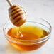 A close up of a honey dipper dripping into a bowl of liquid honey.