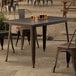 A Lancaster Table & Seating copper outdoor table on a stone patio with a brown chair under it.