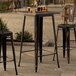 A Lancaster Table & Seating distressed copper bar height table with three black metal chairs on a stone patio.