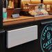 A Regency aluminum folding concession shelf mounted on a white wall above a counter with a tray of pastries and a laptop.