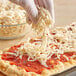 A gloved hand sprinkling BE Hive garlic cheese shreds on a pizza.