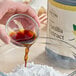 A hand pouring brown Hometown Provisions Gourmet Pure Vanilla Extract into a glass bowl of white powder.