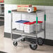 A Regency stainless steel utility cart with two shelves holding bins and a box.