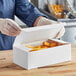 A person in gloves opening a white take-out box of chicken and fries.