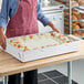 A woman wearing an apron holds a white rectangular tray of frosted cake.