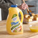 A hand pours Phase garlic butter-flavored oil from a plastic bottle onto yellow liquid in a bowl on a wooden table.