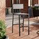 A black Lancaster Table & Seating outdoor bar stool with a gray faux teak seat on a brick patio.