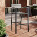 A Lancaster Table & Seating Axel Series outdoor barstool with a black frame and gray faux teak slats on a brick patio.