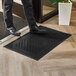 A black 2' x 3' diamond-patterned rubber entrance mat placed on a wooden floor near a doorway.