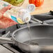 A hand pouring LouAna Liquid Blend Coconut Oil from a plastic bottle into a frying pan on a kitchen counter.