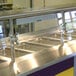 An Advance Tabco cafeteria food shield with a stainless steel shelf on a food counter.