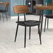 A Lancaster Table & Seating mid-century wooden chair with a black vinyl seat and wood backrest at a restaurant table.