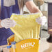 A person in gloves using a Heinz Gold plastic countertop pump to dispense yellow mustard into a bag of food.